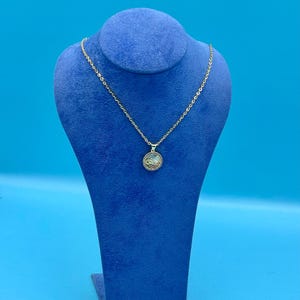 Op de afbeelding: Een gouden ketting met een klein, rond hangertje met een witte steen omringd door kleinere stenen. De ketting wordt gepresenteerd op een blauwe fluwelen standaard.