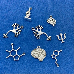 May include: Silver charms featuring science and psychology symbols. The collection includes a microscope, brain, neuron, and a psi symbol. The charms are displayed on a blue background.