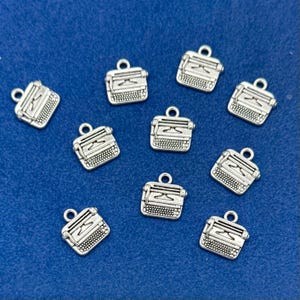 May include: A collection of nine silver-toned metal typewriter charms, each featuring a small loop at the top for attachment. The charms display detailed keys, a carriage, and a platen, all rendered in a metallic finish with dark accents.