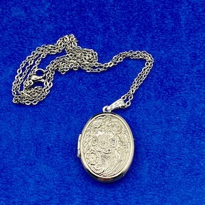 May include: A silver oval locket necklace with a floral design. The locket has a hinged opening and a chain attached to the top.