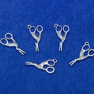 Può includere: Cinque ciondoli a forma di forbici in metallo argentato con un design decorativo. I ciondoli sono a forma di forbici con un motivo dettagliato sulle lame.