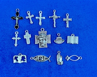 May include: A collection of silver-toned religious charms, including crosses, a fish symbol with "JESUS" inscribed, an open book, and a praying hands charm. The charms are displayed on a blue surface.