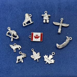 May include: A collection of ten silver-toned metal charms featuring Canadian-themed designs including an ice skate, an inukshuk, a totem pole, a hockey player, a canoe, a wolf, a moose, two maple leaves, and a rectangular Canadian flag charm.