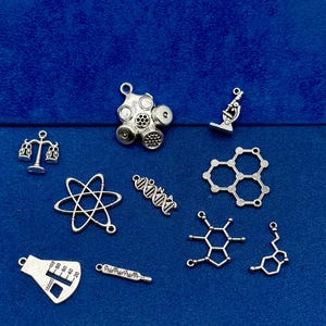 May include: A selection of silver science-themed charms. The charms include a gas mask, microscope, molecular structures, a balance scale, an atom model, a DNA strand, a flask, and a thermometer. The charms are displayed on a blue background.