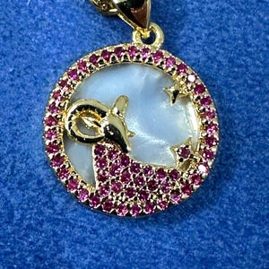 Puede incluir: Collar de oro con un colgante incrustado de cristales rosas en forma de círculo de madreperla blanca con una silueta de carnero dorada.