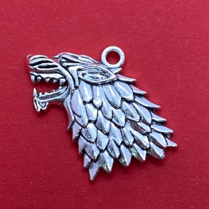 Op de afbeelding: Zilverkleurige metalen hanger met een gedetailleerd wolfskop met een gestructureerd, schubbig ontwerp. De hanger heeft een lus aan de bovenkant om aan een ketting of armband te bevestigen.