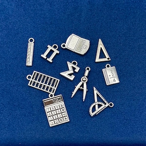 May include: A selection of silver-toned mathematical charms, including a calculator, laptop, abacus, ruler, and geometric shapes. The charms are presented on a dark blue surface.