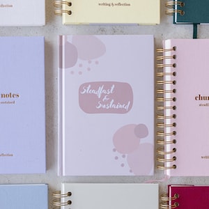 Puede incluir: Una colecci&oacute;n de cuadernos con espiral en varios colores pastel. El cuaderno central es de color rosa claro con las palabras "Steadfast & Sustained" en una fuente suave y manuscrita. Otros cuadernos incluyen "notes" y "chunk" en letras doradas.