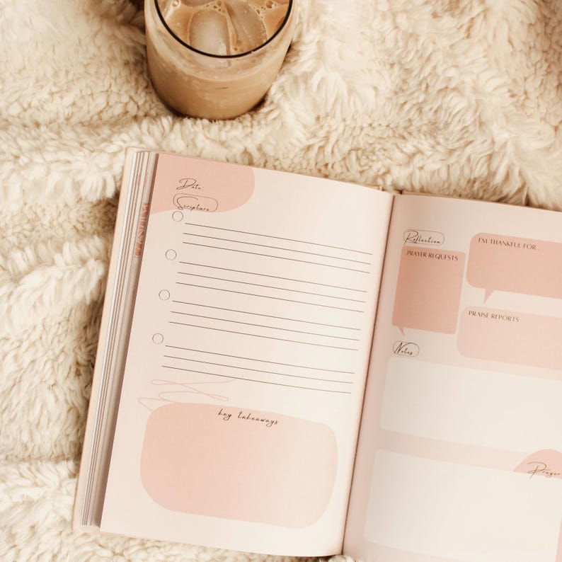 Puede incluir: Diario abierto con detalles en rosa y p&aacute;ginas rayadas. El diario incluye secciones para la fecha, la escritura, las reflexiones, las peticiones de oraci&oacute;n y las notas. Un vaso de caf&eacute; helado est&aacute; en el fondo sobre una manta cremosa y esponjosa.