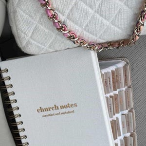May include: A white notebook with gold lettering reading "church notes" and "steadfast and sustained". The notebook is open, showing tabbed pages. A white quilted handbag with a pink and gold chain is visible in the background.
