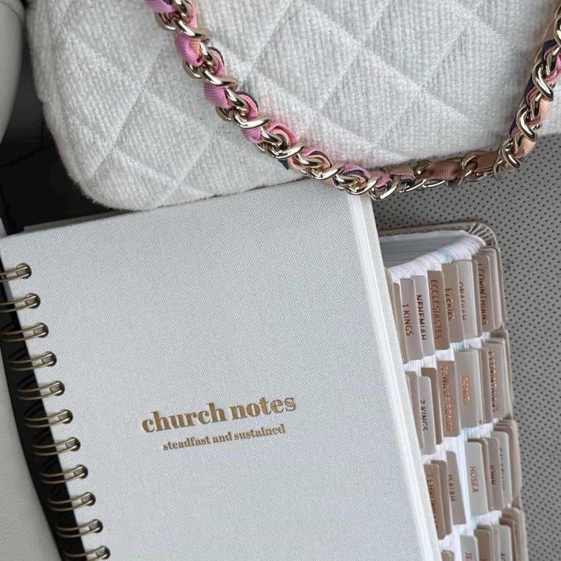 May include: A white notebook with gold lettering reading "church notes" and "steadfast and sustained". The notebook is open, showing tabbed pages. A white quilted handbag with a pink and gold chain is visible in the background.