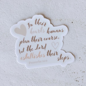 Puede incluir: Una pegatina blanca con un borde marrón claro y texto. El texto dice: "En sus corazones los humanos planean su curso, pero el Señor establece sus pasos. Proverbios 16:9." Hay dos gráficos de corazón.