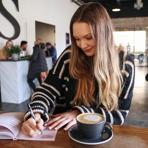 Puede incluir: Una persona escribe en un cuaderno en una mesa con caf&eacute;. Lleva un su&eacute;ter negro con rayas blancas y beige. El caf&eacute; est&aacute; en una taza gris oscuro sobre un platillo a juego.
