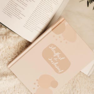 Puede incluir: Un diario de color rosa claro con las palabras "Steadfast & Sustained" en escritura blanca. El diario est&aacute; rodeado de libros abiertos con texto impreso. El fondo es una superficie blanca y esponjosa.
