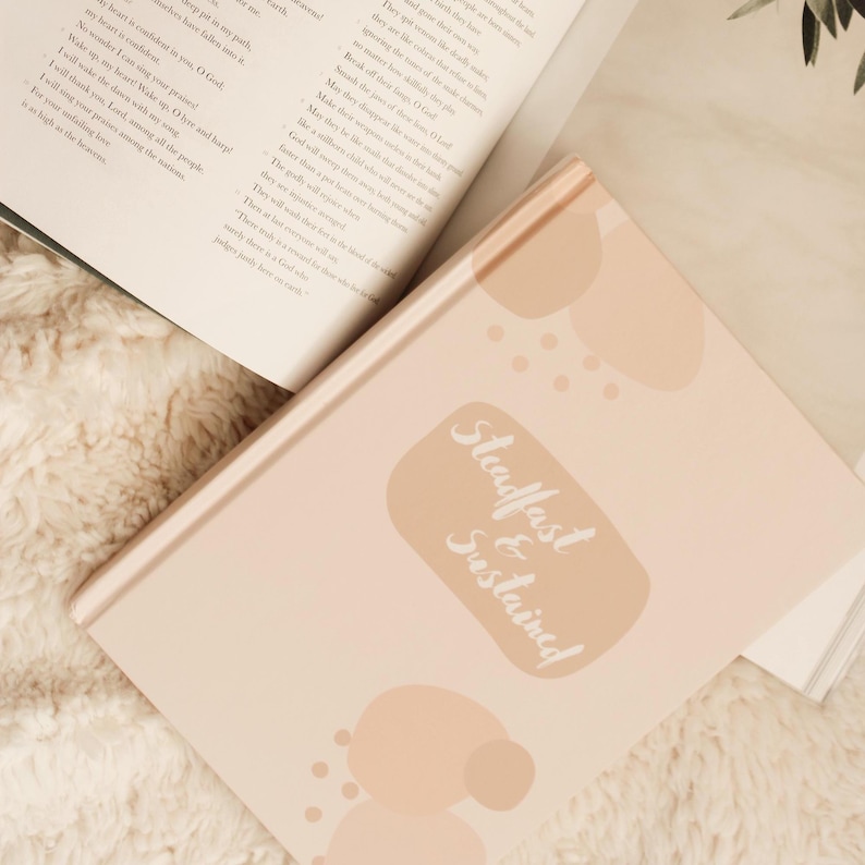Puede incluir: Un diario de color rosa claro con las palabras "Steadfast & Sustained" en escritura blanca. El diario est&aacute; rodeado de libros abiertos con texto impreso. El fondo es una superficie blanca y esponjosa.