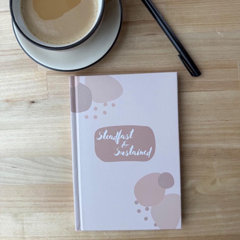 Puede incluir: Un diario de tapa dura con las palabras "Steadfast & Sustained" en un &oacute;valo rosa suave. El diario tiene una cubierta blanca con formas y puntos rosas abstractos. Una taza de caf&eacute; y un bol&iacute;grafo tambi&eacute;n est&aacute;n sobre la superficie de madera.
