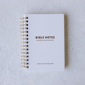 May include: A white spiral-bound notebook with gold-toned wire binding. The cover features dark brown text that reads BIBLE NOTES, steadfast and sustained, and daily devotional guide. The notebook has a textured fabric-like cover material.