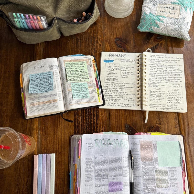 May include: A wooden table displays an open Bible marked with the word "Romans", alongside notebooks, pens, and highlighters. A canvas bag and a small cushion with the text "I need thee every hour" are also visible, creating a scene of study and reflection.