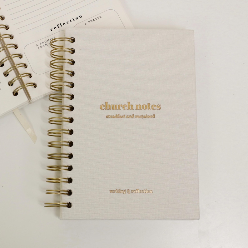 May include: Cream-coloured spiral-bound notebook with gold lettering that reads "church notes" and "writing & reflection." The notebook is open to a page with lines and the word "reflection" printed at the top.