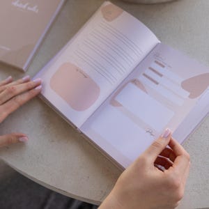 Puede incluir: Diario abierto con p&aacute;ginas rayadas y detalles en rosa suave. El diario est&aacute; abierto por las manos de una persona, revelando un dise&ntilde;o con espacio para notas e indicaciones. La portada de un segundo diario es visible en el fondo.