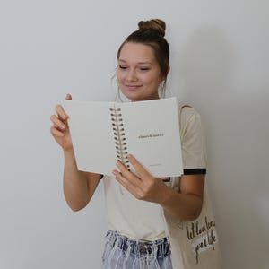May include: A cream-coloured spiral-bound notebook with the words "church notes" printed on the open page. The notebook has a gold spiral binding. The person is holding the notebook and looking at it.