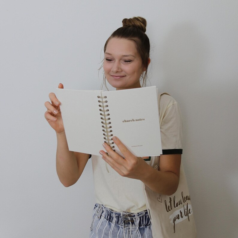 May include: A cream-coloured spiral-bound notebook with the words "church notes" printed on the open page. The notebook has a gold spiral binding. The person is holding the notebook and looking at it.