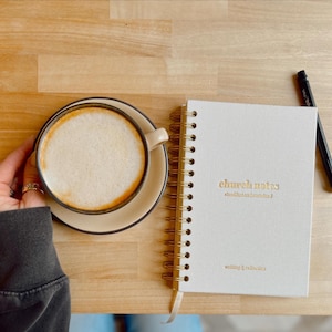 May include: A coffee cup with a creamy top sits on a saucer beside a spiral-bound notebook. The notebook is white with gold lettering that reads "church notes" and "writing & reflection". A black pen rests on the wooden surface.