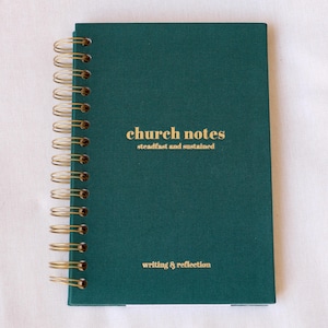 May include: A dark teal spiral-bound notebook with gold lettering that reads "church notes steadfast and sustained" and "writing & reflection." The spiral binding is gold. The notebook is on a white surface.