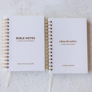 May include: Two white spiral-bound notebooks with gold coils. One notebook reads "BIBLE NOTES" and "daily devotional guide." The other reads "church notes" and "writing & reflection." Both notebooks have the phrase "steadfast and sustained."