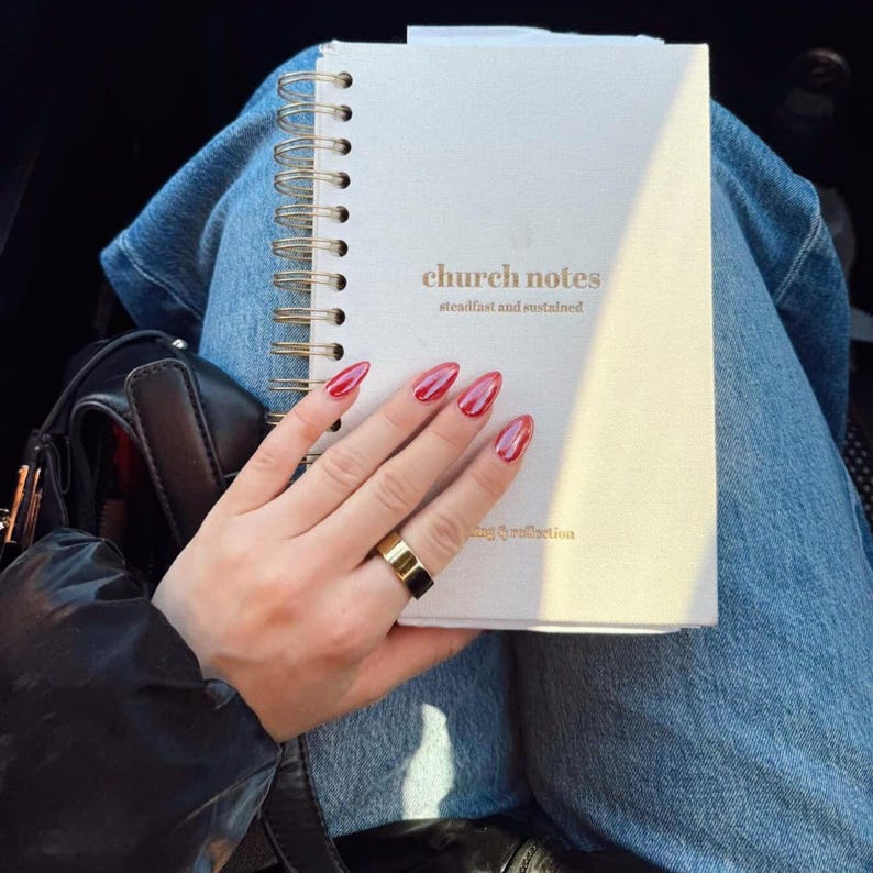 May include: A cream-coloured spiral-bound notebook with the words "church notes" in gold. The notebook is held by a hand with red nail polish and a gold ring. The text "steadfast and sustained" is below the title.