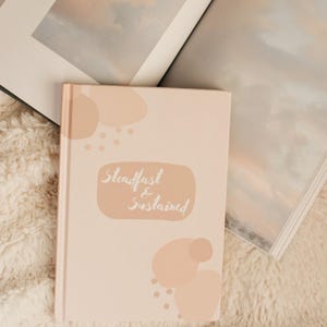 Puede incluir: Un diario de color rosa claro con las palabras "Steadfast & Sustained" en escritura cursiva. El diario tiene una cubierta blanda con formas abstractas de color melocot&oacute;n. Dos libros abiertos en el fondo, uno con una imagen de paisaje.