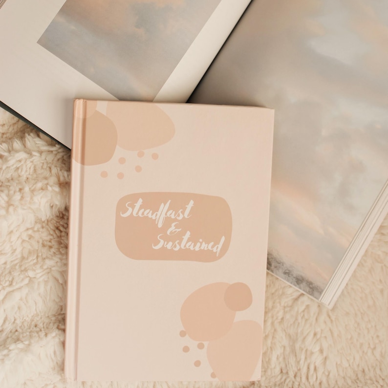 Puede incluir: Un diario de color rosa claro con las palabras "Steadfast & Sustained" en escritura cursiva. El diario tiene una cubierta blanda con formas abstractas de color melocot&oacute;n. Dos libros abiertos en el fondo, uno con una imagen de paisaje.