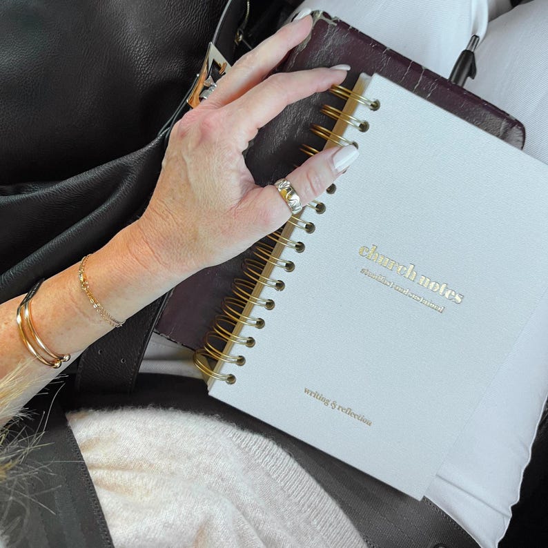 May include: A white spiral-bound notebook with gold lettering that reads "church notes" and "writing & reflection." The notebook is held open, revealing the gold spiral binding. A dark leather notebook and black bag are in the background.
