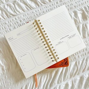 May include: A cream-coloured, spiral-bound journal with sections for notes, scripture, and reflection. It has a gold spiral binding and is placed on top of an orange notebook with a floral design. The journal is open, revealing lined pages.