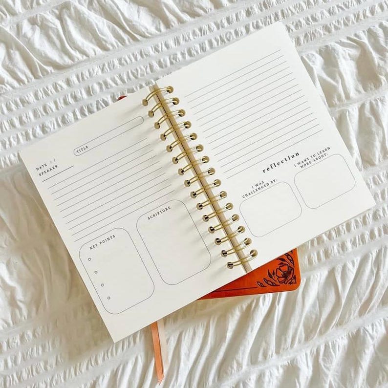 May include: A cream-coloured, spiral-bound journal with sections for notes, scripture, and reflection. It has a gold spiral binding and is placed on top of an orange notebook with a floral design. The journal is open, revealing lined pages.