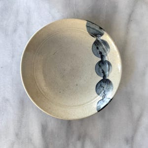 May include: A round ceramic plate with a speckled cream-colored glaze. The plate features a vertical row of five dark grey, circular brushstroke patterns along the right side, with a smooth, slightly concave center and a raised outer rim.