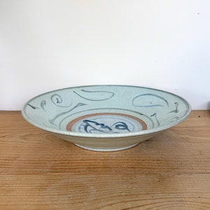 May include: A handmade ceramic bowl with a light blue glaze and decorative blue and brown painted details. The bowl features a wide, shallow design, perfect for serving or display. The interior has a central design.