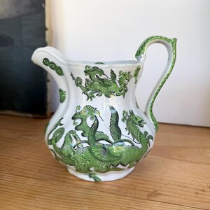 May include: A white ceramic creamer pitcher featuring a repeating green dragon pattern. The pitcher has a curved handle, a scalloped rim, and a pouring spout. The green dragon design is detailed with scales and wings, wrapping around the body.