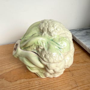 May include: A ceramic cauliflower-shaped lidded container featuring a realistic textured surface with cream and pale green glazed coloring. The vegetable-themed vessel includes a small square label on the side with a dark graphic design.