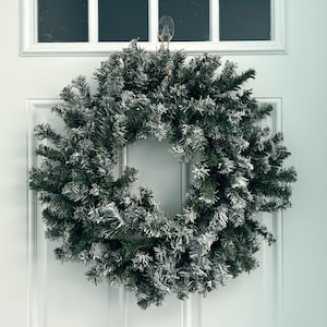 May include: A green artificial Christmas wreath with a snowy, frosted appearance. The wreath is circular and hangs on a white door. The wreath is made of evergreen branches and is a festive holiday decoration.
