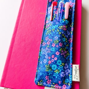 Puede incluir: Un porta bolígrafos de tela azul con estampado floral y una banda elástica. El porta bolígrafos está unido a una libreta rosa fuerte con el texto "2TreeRun" en el lateral.