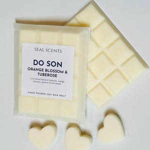 May include: A white soy wax melt in a clear plastic package with the text "SEAL SCENTS DO SON ORANGE BLOSSOM & TUBEROSE" and "HAND POURED SOY WAX MELT". The wax melt is shaped like a bar and is divided into squares. There are three heart-shaped wax melts on a white surface.