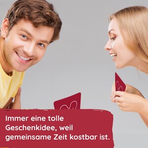 May include: Two people are playing a card game. The cards are red and have a white silhouette of two people on them. The text on the bottom of the image reads "Immer eine tolle Geschenkidee, weil gemeinsame Zeit kostbar ist."