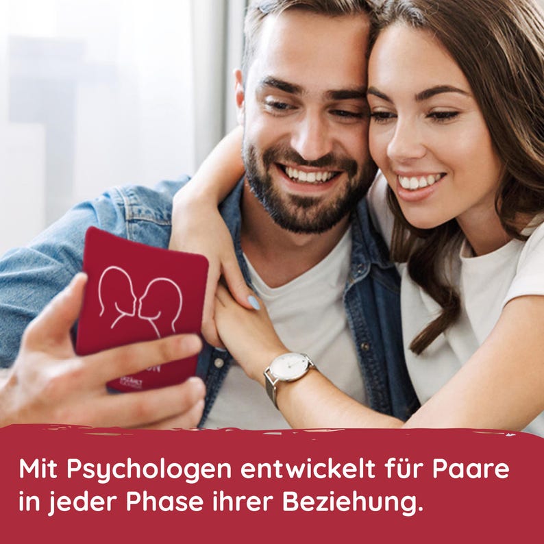 May include: A couple smiles while looking at a red card game with a white outline of two people kissing. The text on the card reads "Intimacy". The text below the image reads "Mit Psychologen entwickelt f&uuml;r Paare in jeder Phase ihrer Beziehung."