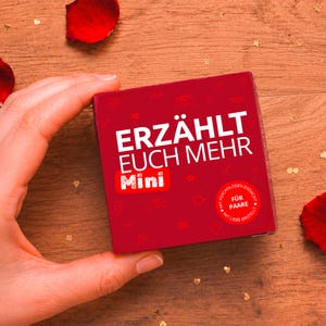 Könnte beinhalten: Eine quadratische rote Schachtel mit dem weißen Text ERZÄHLT EUCH MEHR und Mini in einem roten Kasten. Die Oberfläche ist mit kleinen weißen Strichzeichnungen von Herzen, Augen und Lippen verziert. Ein weißes Siegel trägt den Text MIT PSYCHOLOGEN ERDACHT FÜR PAARE.