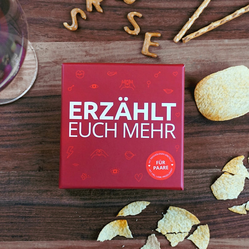 May include: Red box with the text "ERZ&Auml;HLT EUCH MEHR" in white. The box has a smaller red circle with the text "F&Uuml;R PAARE" in white. The box is on a wooden surface with some broken potato chips and pretzels.