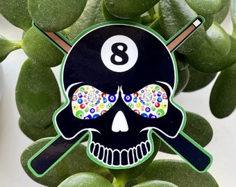 May include: Black and white skull sticker with green outline. The skull's eyes are made up of a pattern of colorful billiard balls. The skull is wearing a black billiard ball with the number 8 on it. Two crossed pool cues are behind the skull.