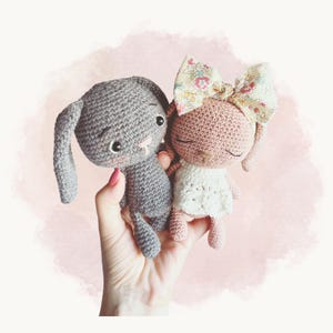 Pode incluir: Dois pequenos animais de peluche feitos de crochê, um coelho cinzento com olhos pretos e um coelho cor-de-rosa com olhos fechados, vestindo um vestido branco e um grande laço com padrão floral na cabeça, ambos com acabamento em fio texturizado.