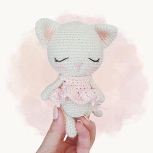 Pode incluir: Uma pequena boneca de gato em crochê na cor branco sujo, com uma saia de babados rosa claro, olhos fechados bordados, um pequeno nariz rosa e um blush rosa sutil nas bochechas. O gato tem orelhas pontudas com detalhes em rosa claro.
