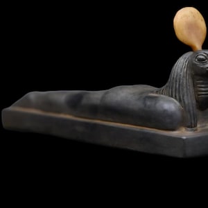 Może przedstawiać: Czarna kamienna statua egipskiego boga Horusa, przedstawiona jako sokół z ludzką głową, leżąca na prostokątnej podstawie. Statua ma żółty kamienny nakrycie głowy.